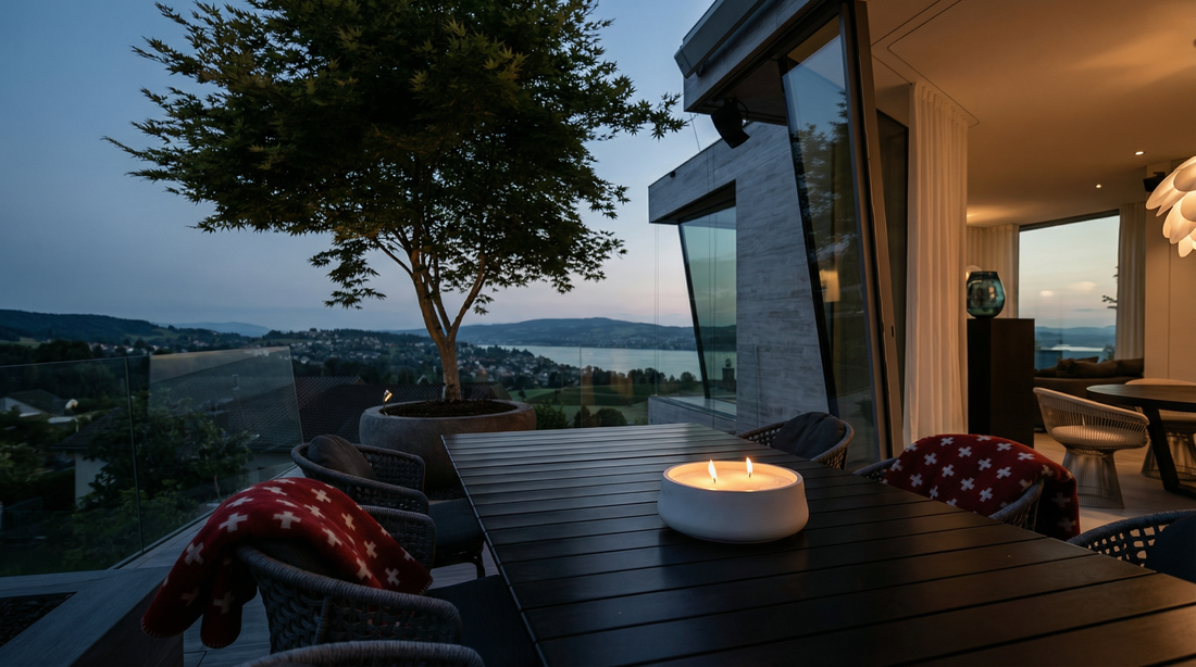 Kerzenschale mit brennender Kerze auf einem Gartentisch auf moderner Terrasse bei sommerlicher Abenddämmerung mit Blick auf Zürichsee und Uetliberg